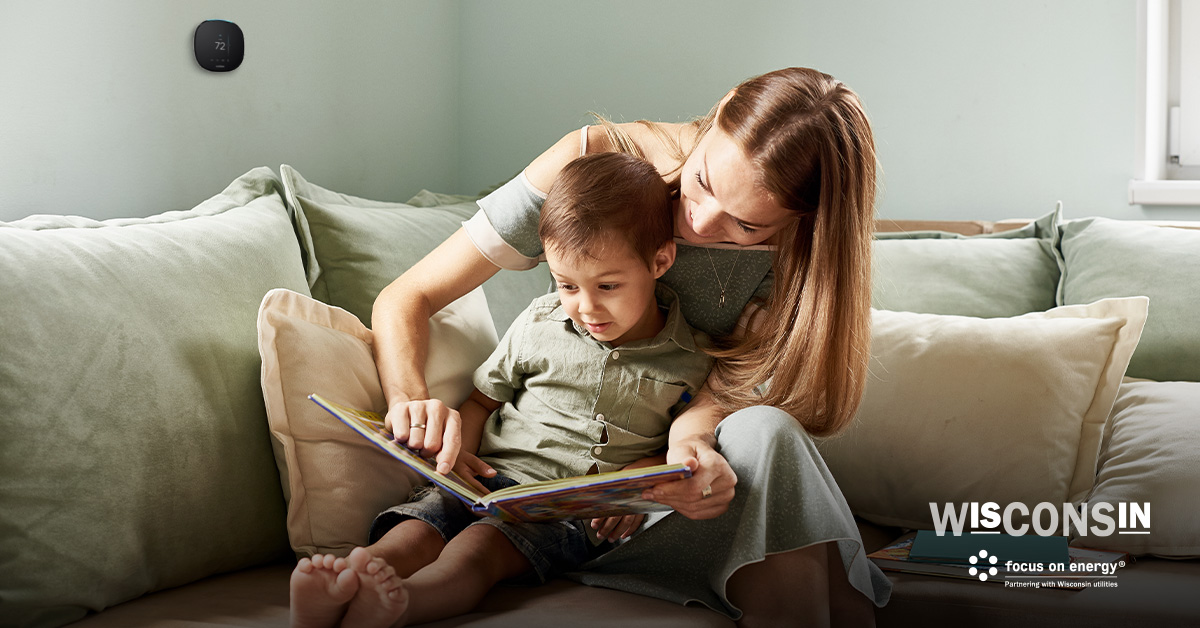 A mother and son read a book together on the couch. A smart thermostat can be seen on the wall in the background.