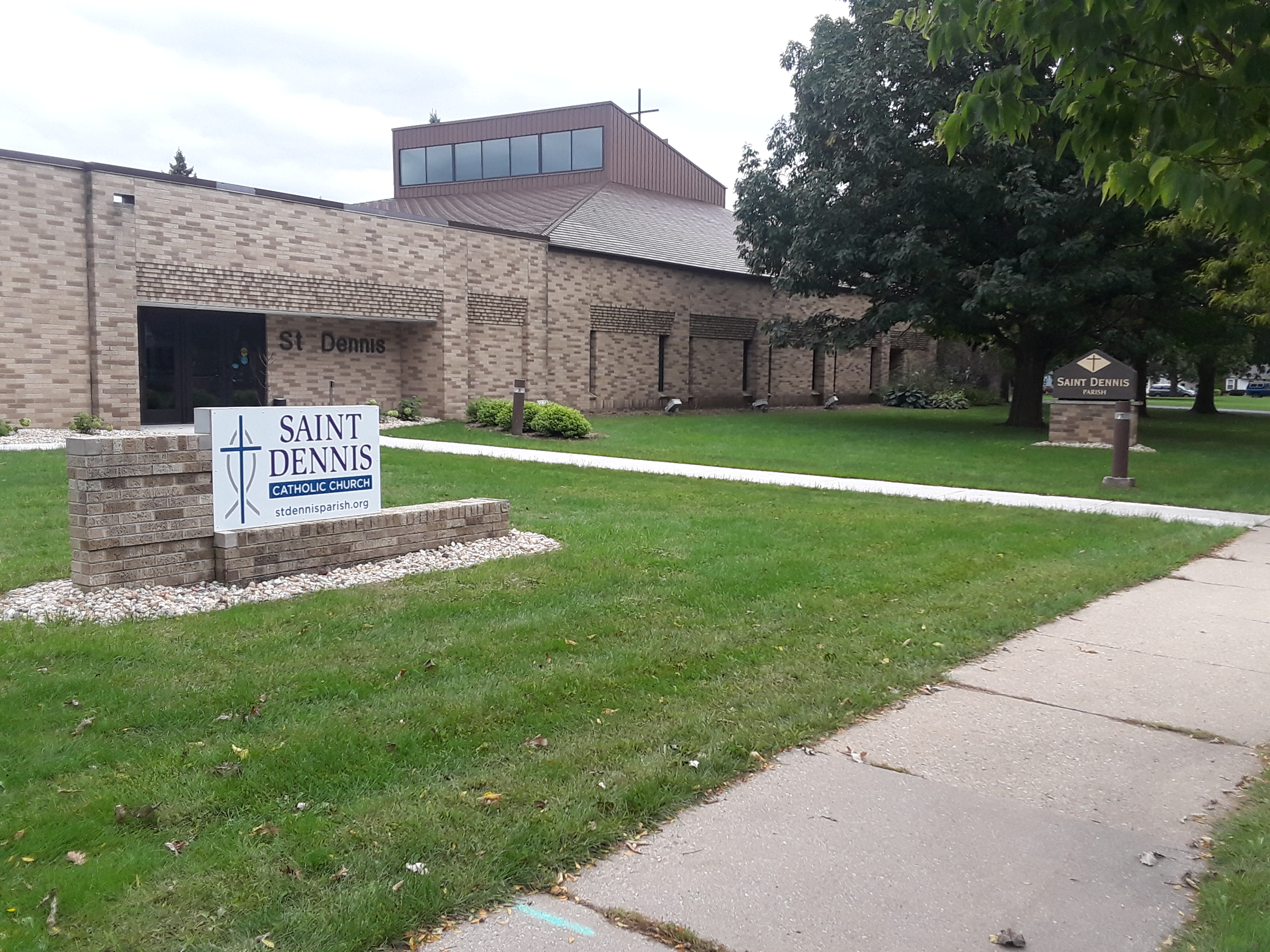 A brown brick building can be seen with a green lawn and trees in front of it, as well as a sidewalk. There is a white sign that reads "Saint Dennis Catholic Church stdennisparish.org" in the front of the building.