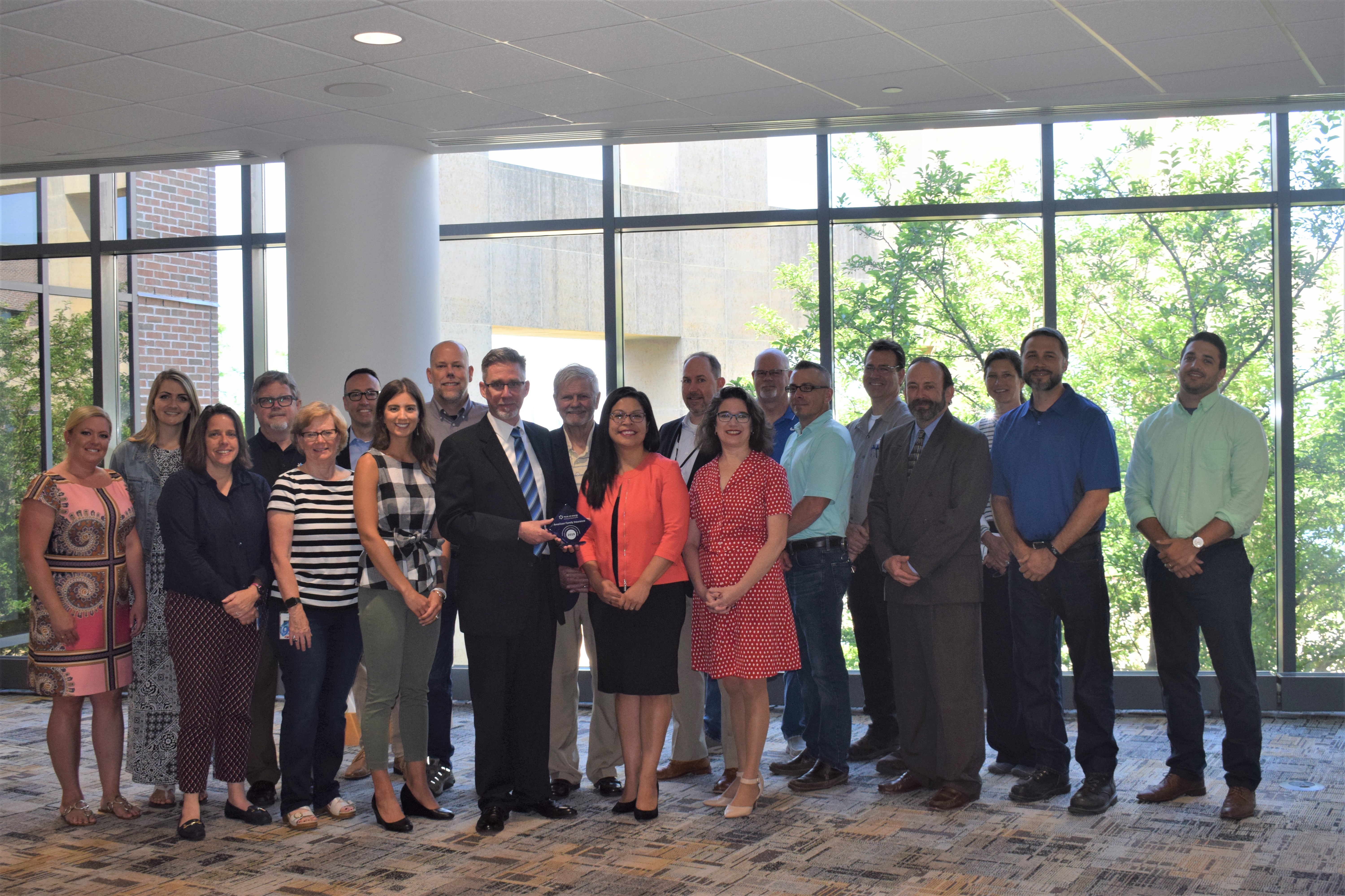 Large group of people stand together in a business building as a man in the center holds the blue award. There are windows with trees and greenery in the background.