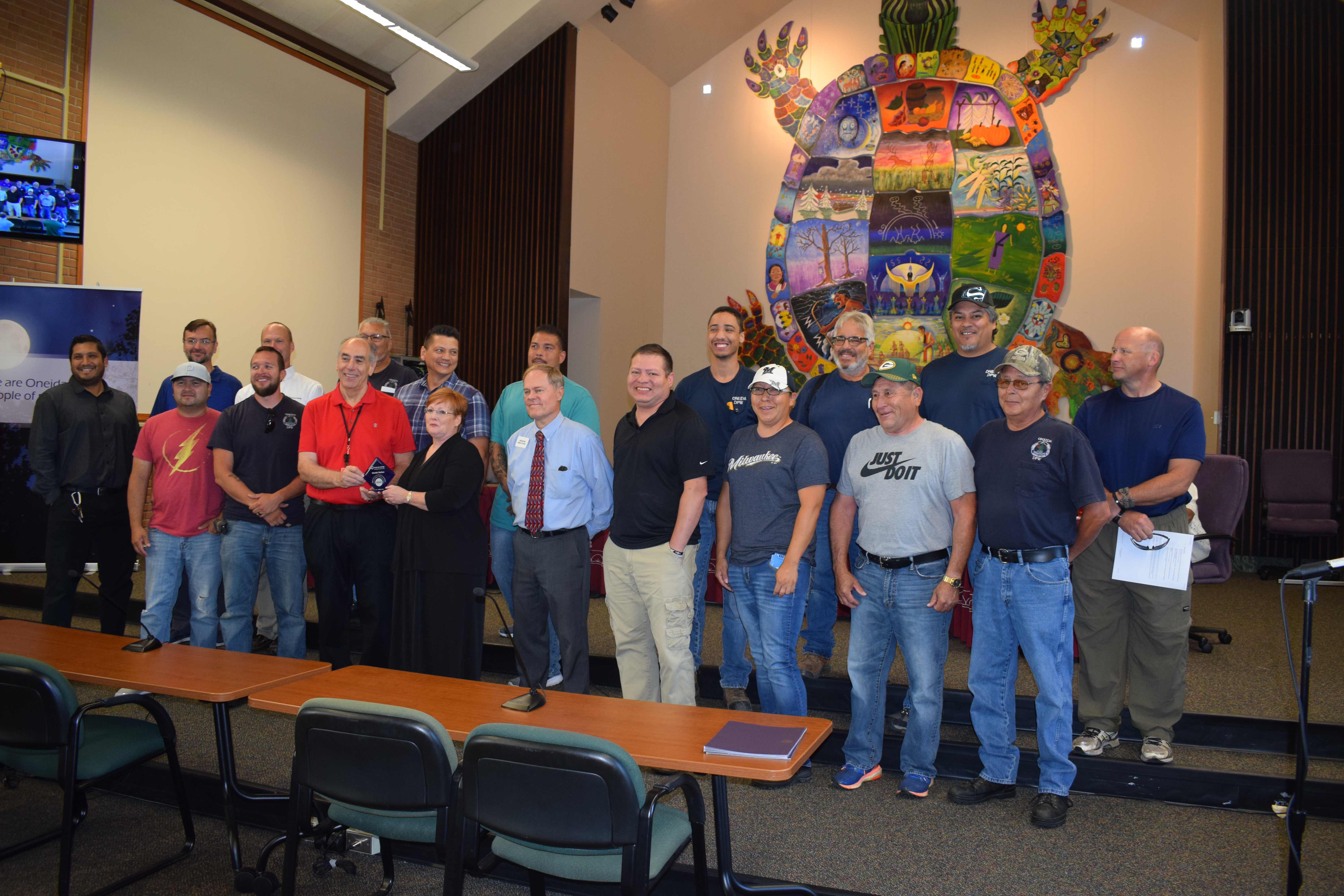 A group of people stand inside a community building. The person in the center holds a blue award.