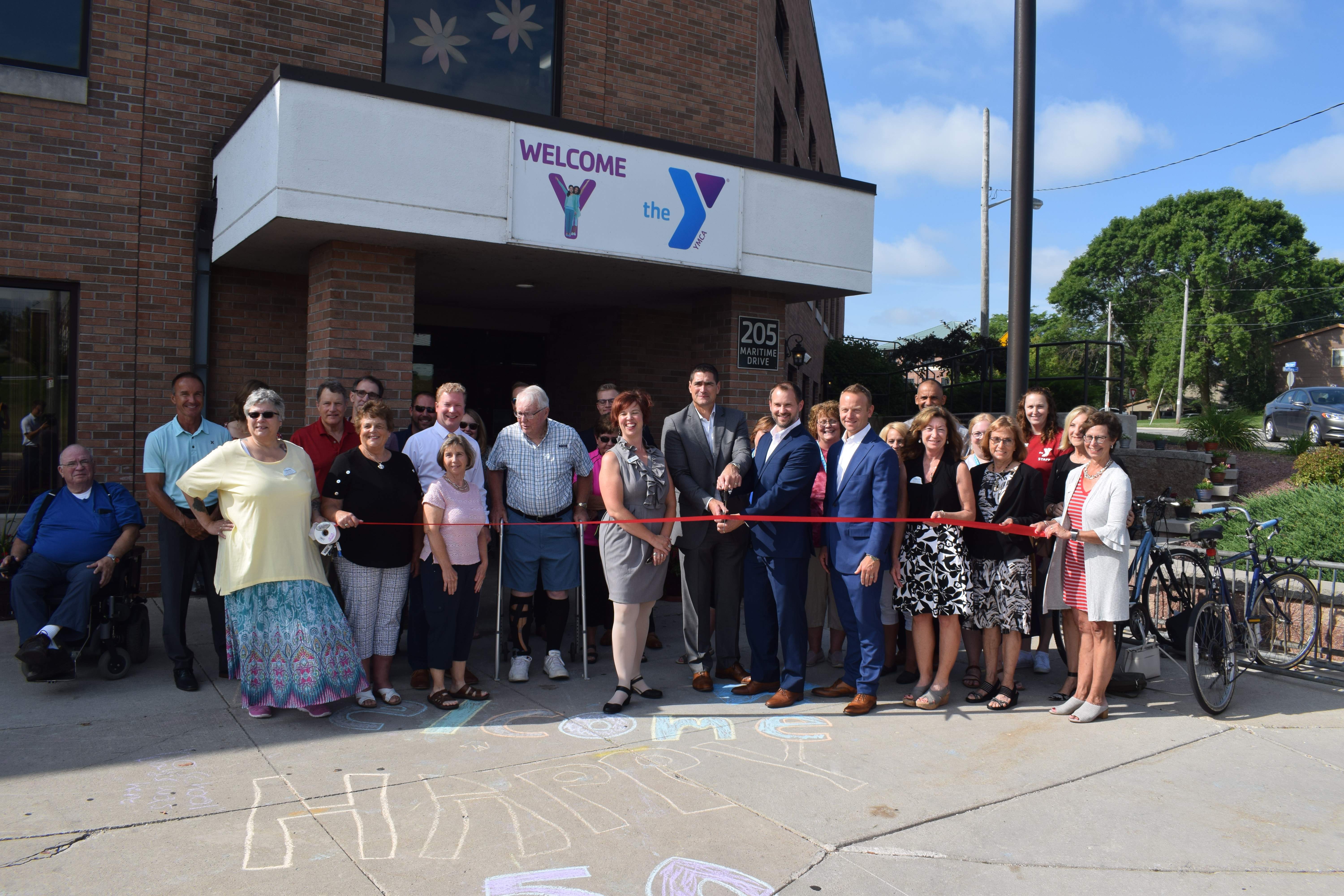 A large group of people are gathered in front of the entrance to a building on a sunny summer day. There is a red ribbon and a man in the center holds scissors to cut it. There is a sign above that says "Welcome" and "the Y".