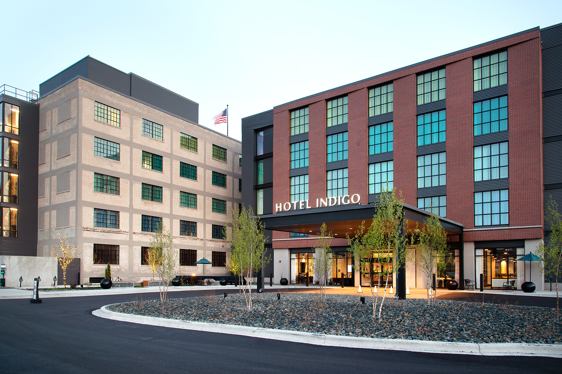 Exterior shot of large, multi-story building with a circular driveway and valet pavilion. The letters "Hotel Indigo" can be seen above the entrance and an American Flag is displayed in front of the building. 