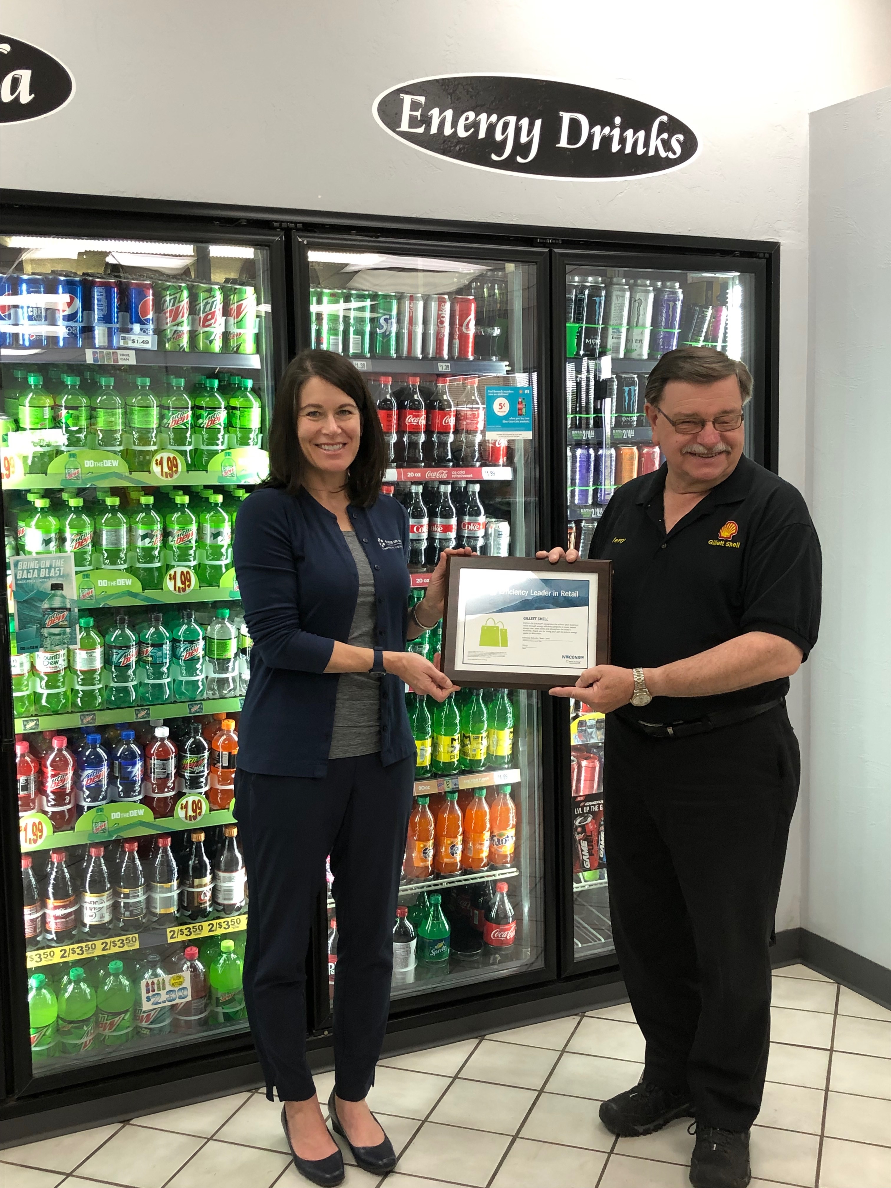 Two people stand in a grocery store, holding a plaque. Behind them is a wall of refrigerators filled with beverages, under a sign that reads "Energy Drinks".