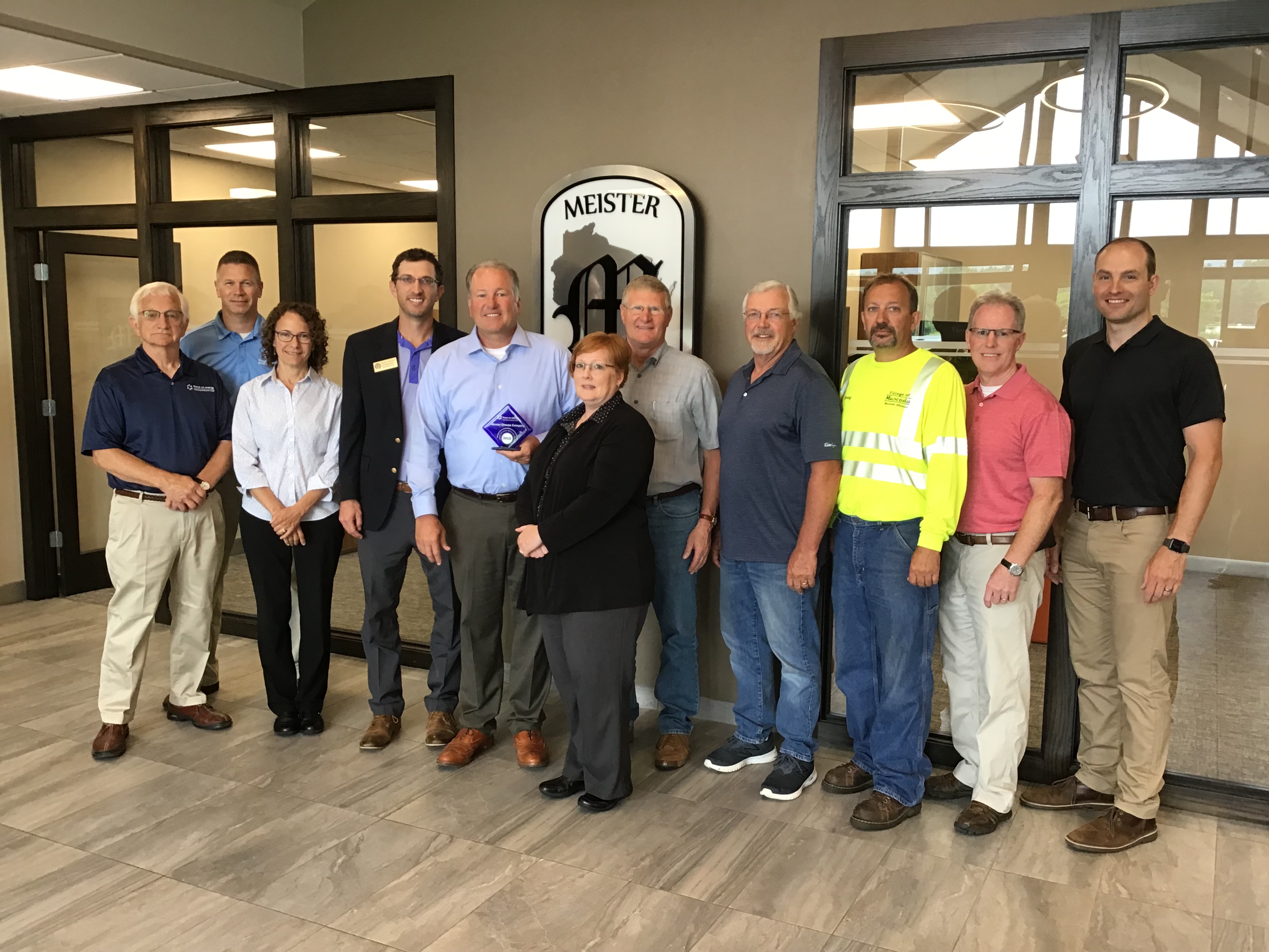 A group of people stand below the Meister Cheese sign. The man in the center of the group holds the blue Focus on Energy Award. 