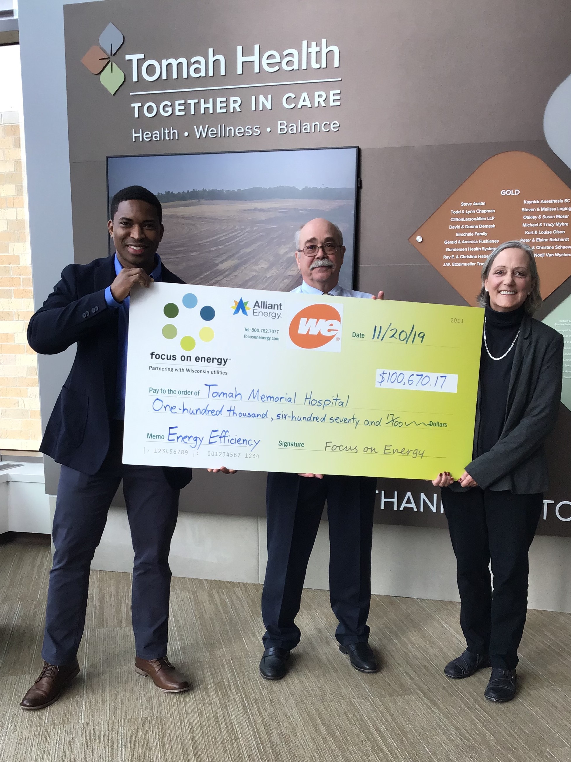 Three people in business attire hold a large, green check in front of a sign that says "Tomah Health" with "Together in Care Health Wellness Balance" written underneath it.