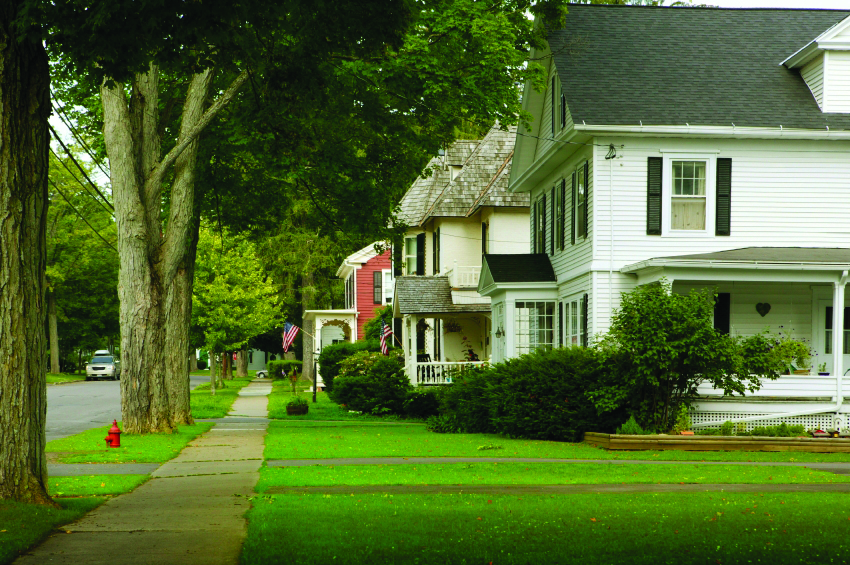 Image of a neighborhood street with one white, two story home featured. There is a large green lawn and a sidewalk with trees to the left. 